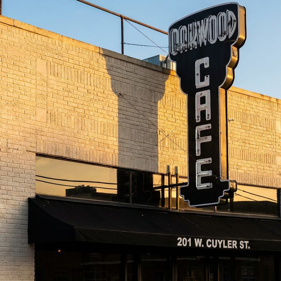 Oakwood Cafe's brick exterior with a prominent vertical sign and black awnings in golden sunlight.